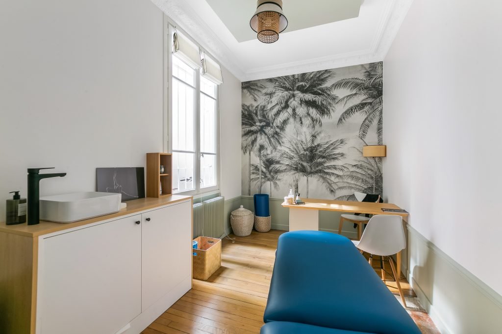 Salle de soins : lavabo et rangements à gauche, grande fenêtre, mur de palmiers, bureau en bois, table de soins bleue au premier plan